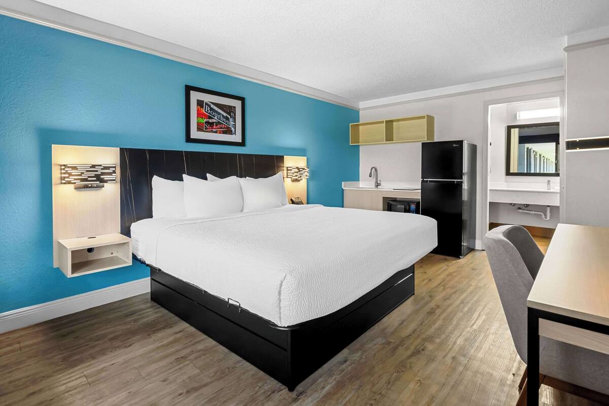 A modern hotel room with a large bed, a desk, a kitchenette, and a bathroom.