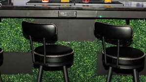 Zona de bar con varias botellas de licor en estantes, tres taburetes y una barra.