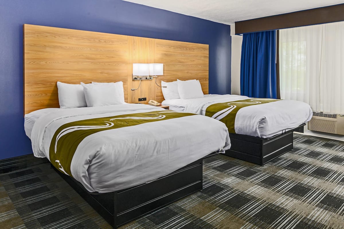 Quarto de hotel com duas camas, uma cabeceira de madeira, uma mesa de cabeceira, um telefone e uma janela com cortinas azuis.