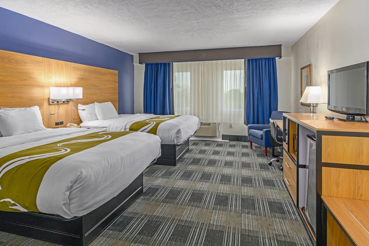 Quarto de hotel com duas camas, uma escrivaninha com televisão, uma cadeira, luminária de mesa e janela com cortinas.