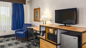 A hotel room with a wooden desk, a television, a microwave, a refrigerator, a blue armchair, a black office chair, a lamp, and a framed picture on the wall. - Quality Inn Bourne - Cape Cod (Buzzards Bay)