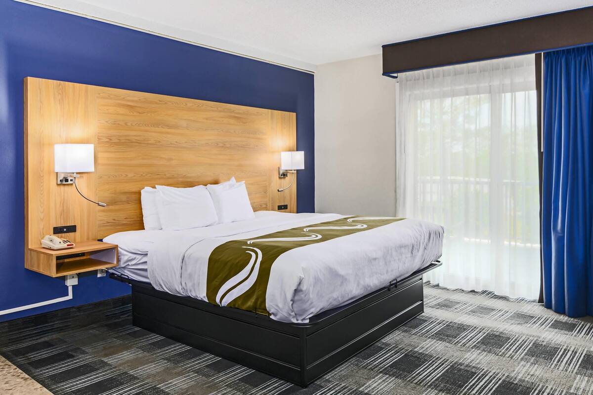Quarto de hotel com uma cama grande, cabeceira de madeira, duas luminárias de apoio, telefone e janela com cortinas azuis.