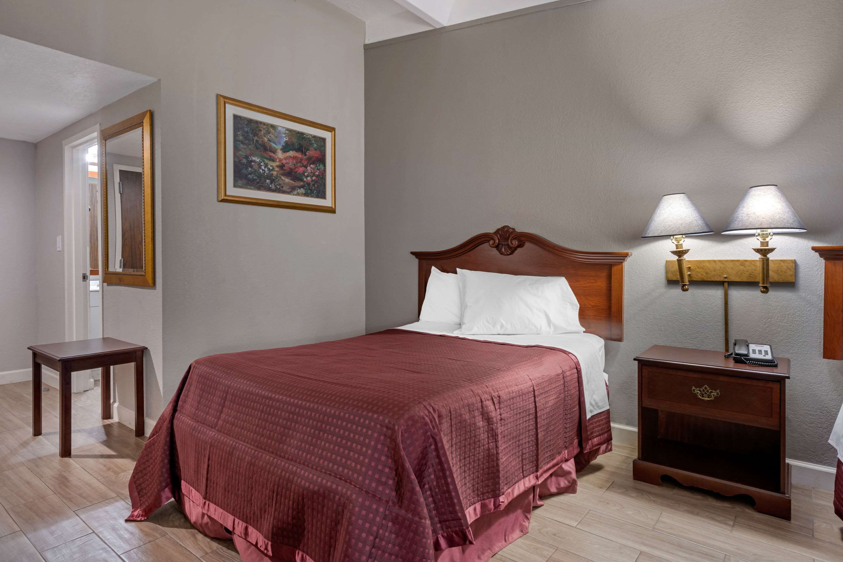 A hotel room with a large bed, a bedside table, a nightstand with a lamp, a mirror, and a framed picture on the wall.