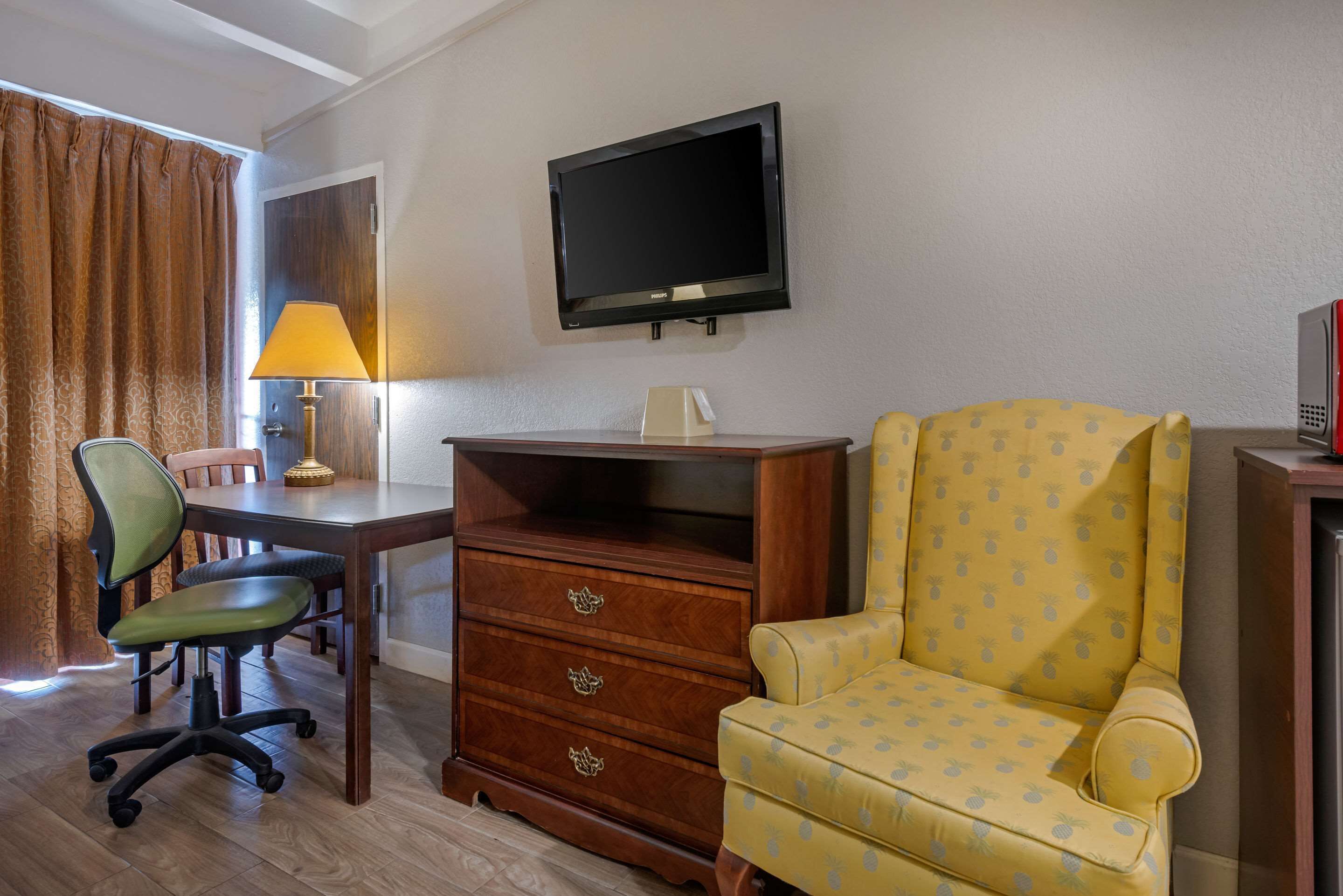 Ein Hotelzimmer mit einem Holztisch, einem Stuhl, einem an der Wand montierten Fernseher und einem gemusterten Sessel.