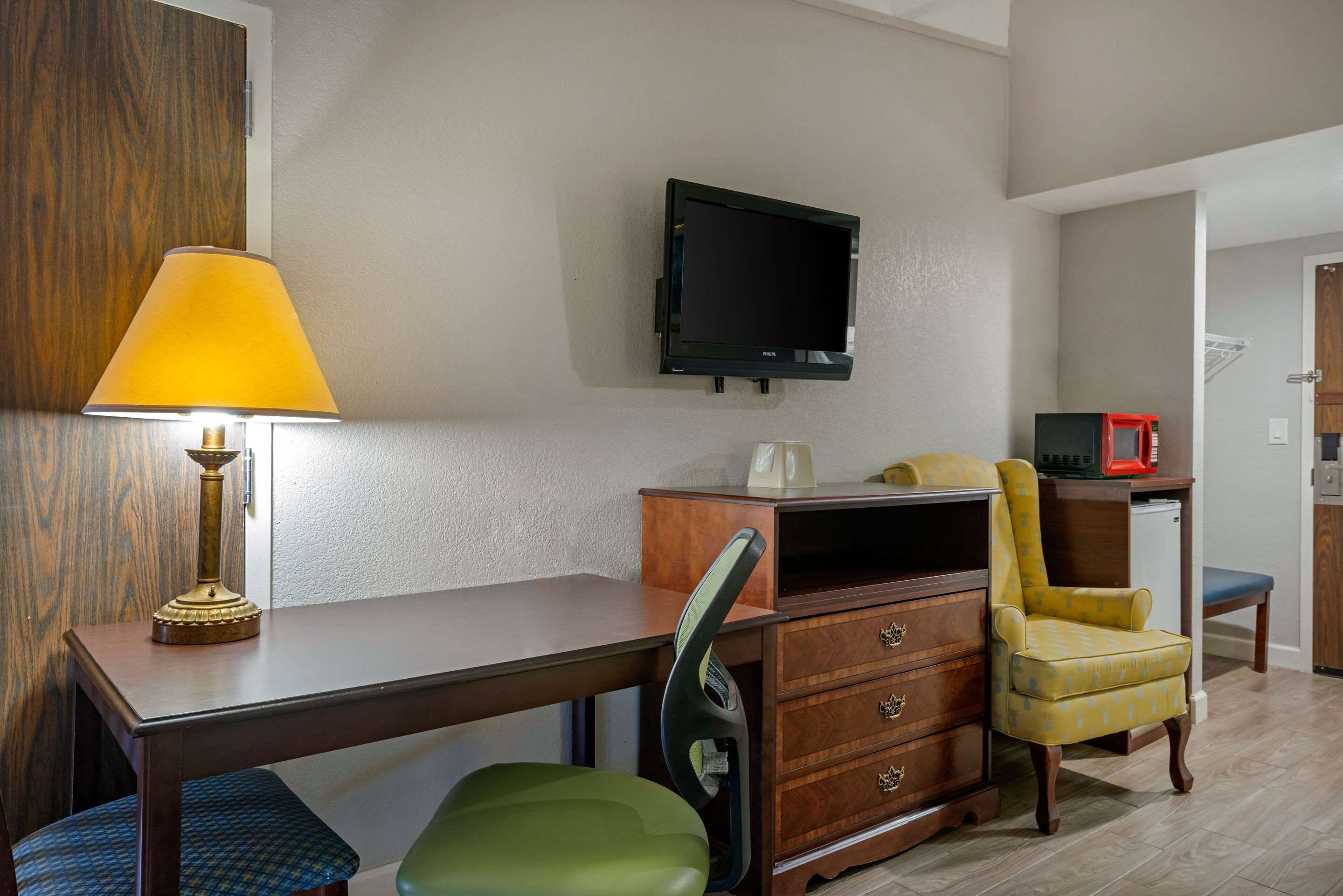 A compact room with a desk, a chair, a television mounted on the wall, and a microwave.