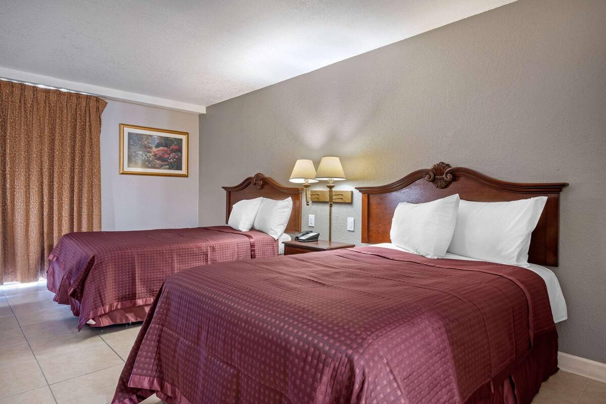 Ein Hotelzimmer mit zwei Betten, einem Nachttisch mit Lampe, einem Bild an der Wand und einem Fenster mit Vorhängen.