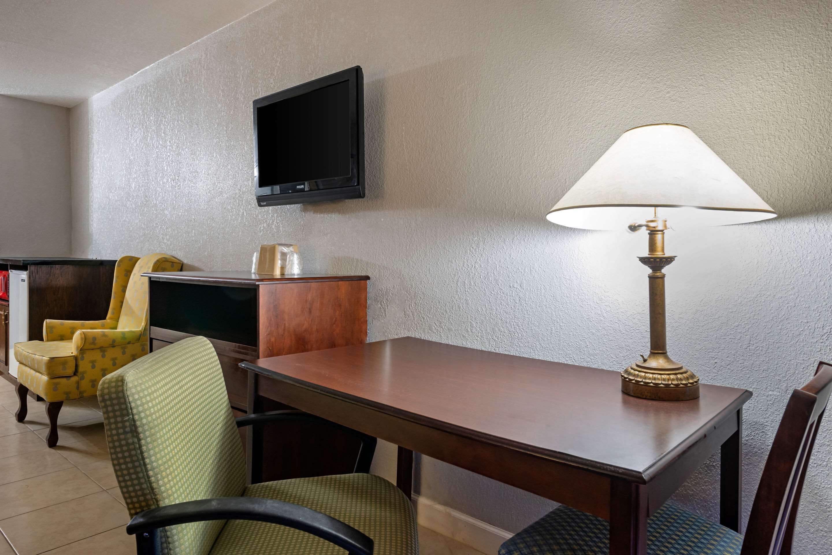 Habitación de hotel con una mesa de comedor de madera, dos sillas, un televisor montado en la pared y una lámpara de mesa.