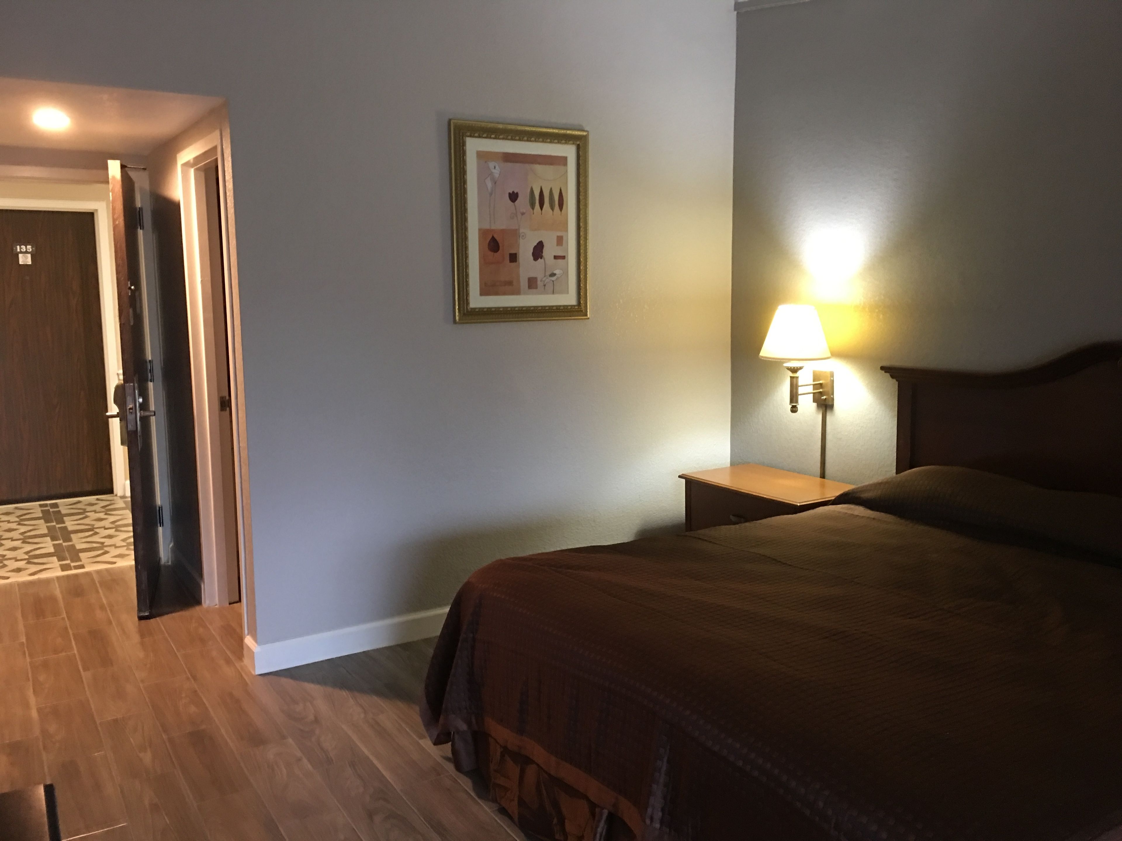 A hotel room with a bed, a nightstand with a lamp, a framed picture on the wall, and a door leading to another room.