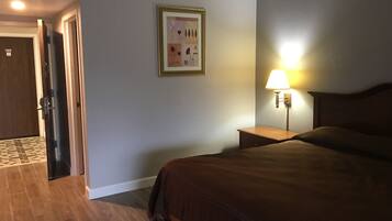 A hotel room with a bed, a nightstand with a lamp, a framed picture on the wall, and a door leading to another room.