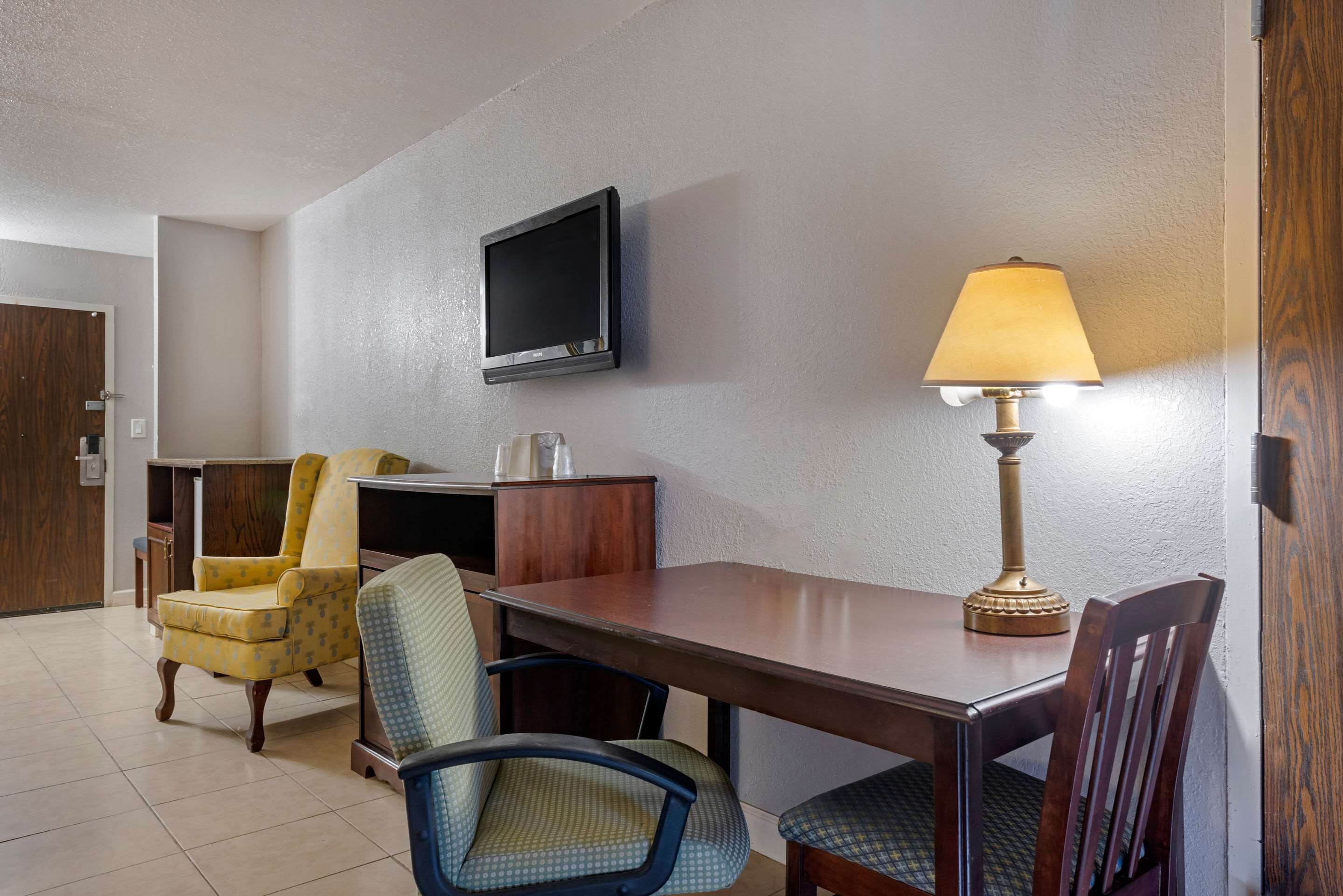 A hotel room with a wooden table, two chairs, a television mounted on the wall, and a table lamp.