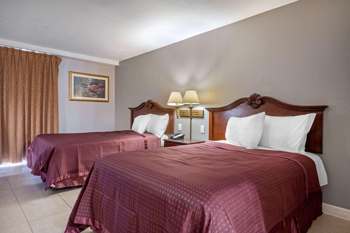 A hotel room with two beds, a nightstand with a lamp, a painting on the wall, and a window with curtains.