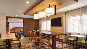 Un comedor de hotel con una gran mesa de madera, varias sillas y una alfombra a rayas de colores. Hay un televisor montado en la pared y una ventana con cortinas.