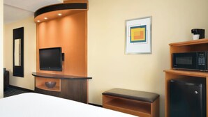 Habitación de hotel con cama, televisión de pantalla plana, microondas, refrigerador, pequeño banco y una obra de arte enmarcada en la pared.