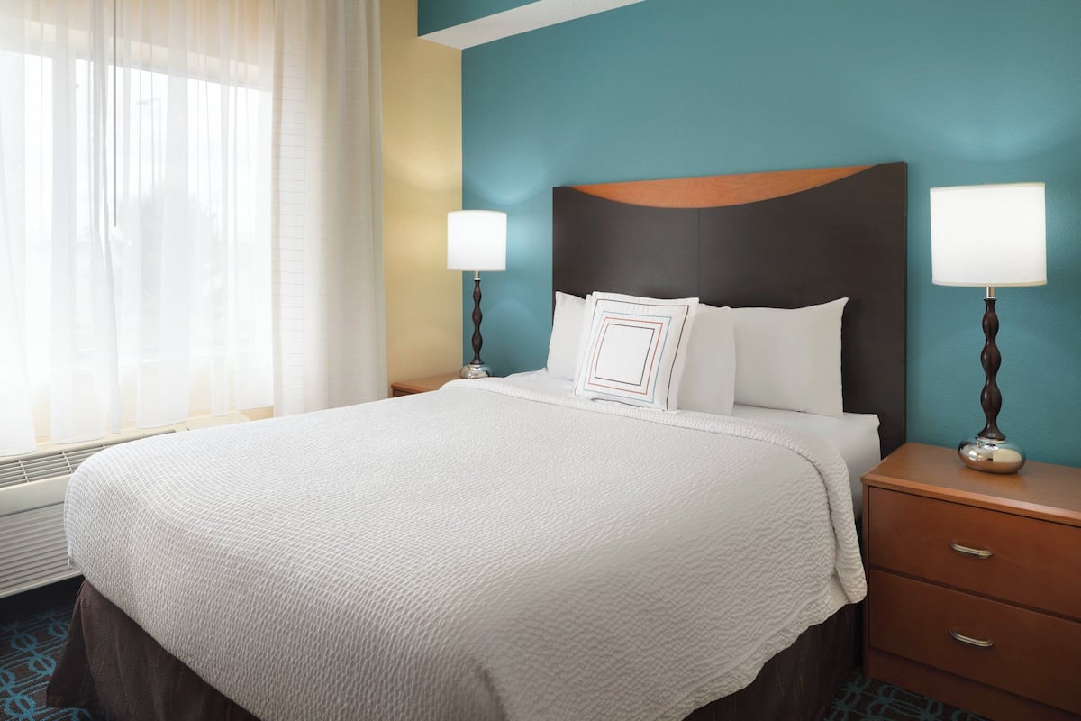 Habitación de hotel con una cama grande, dos mesitas de noche con lámparas, un ventanal con cortinas traslúcidas y una pared de acento en color aguamarina.