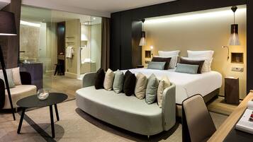 A modern hotel room with a large bed, a sofa, a desk, and a glass-enclosed bathroom.