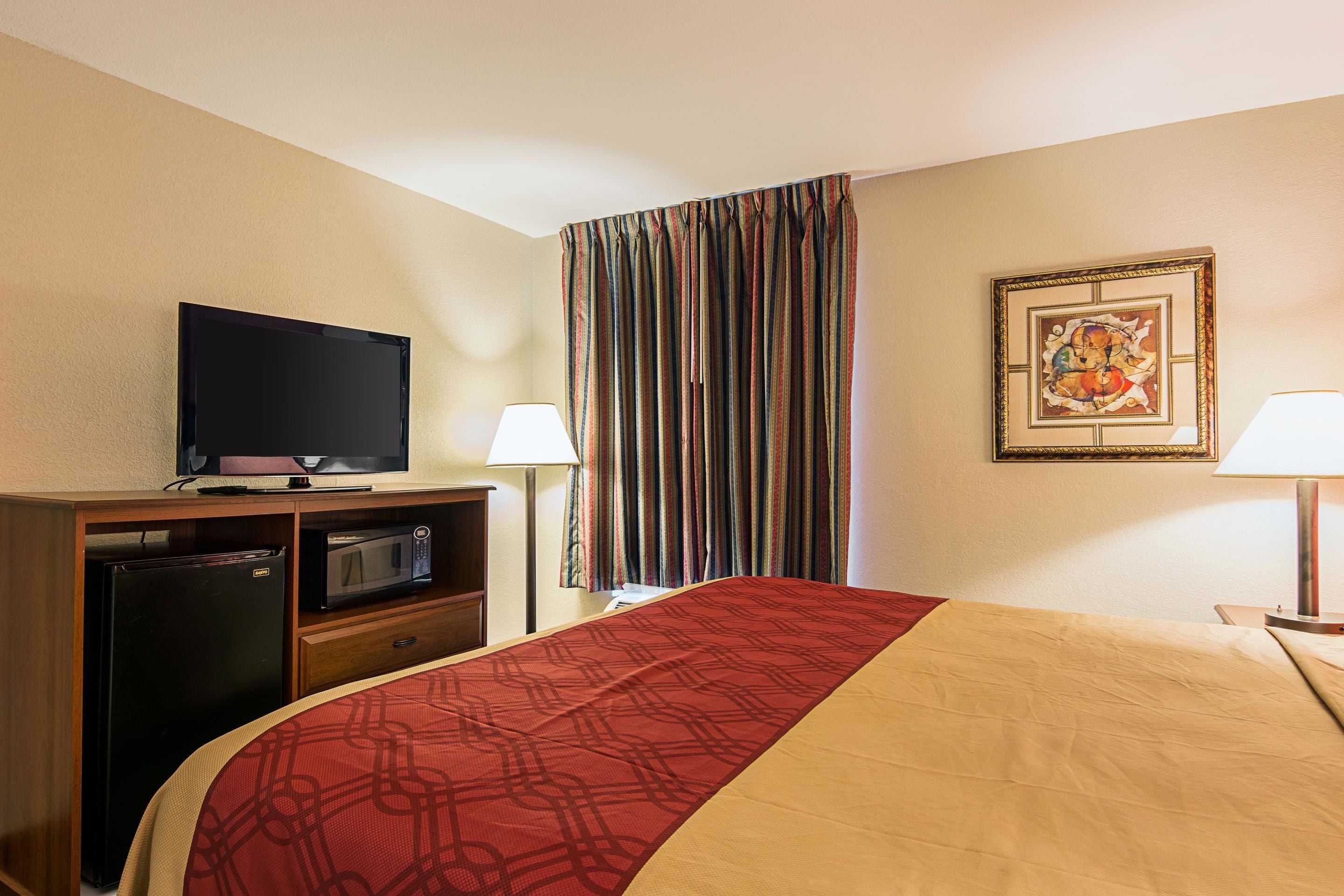 Habitación de hotel con cama, televisor, microondas, refrigerador, lámpara y cortina.