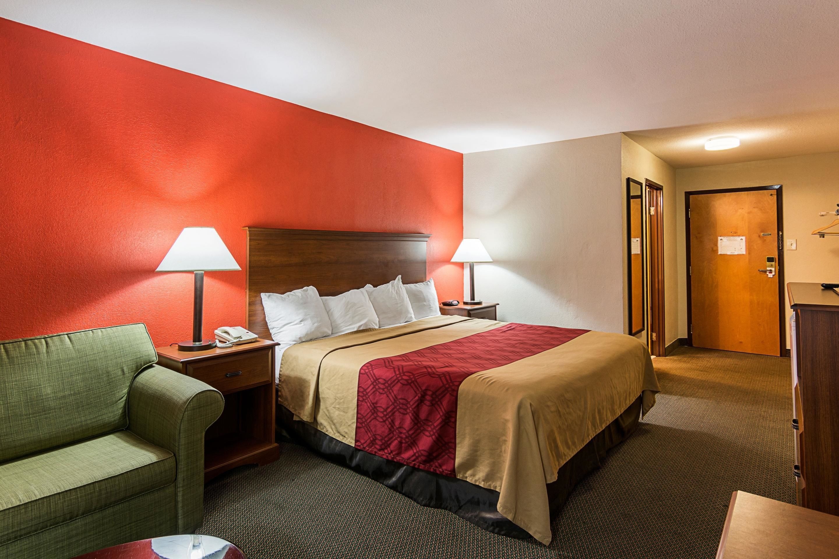 A hotel room with a bed, two bedside lamps, a green armchair, a wooden nightstand, and a wooden door.