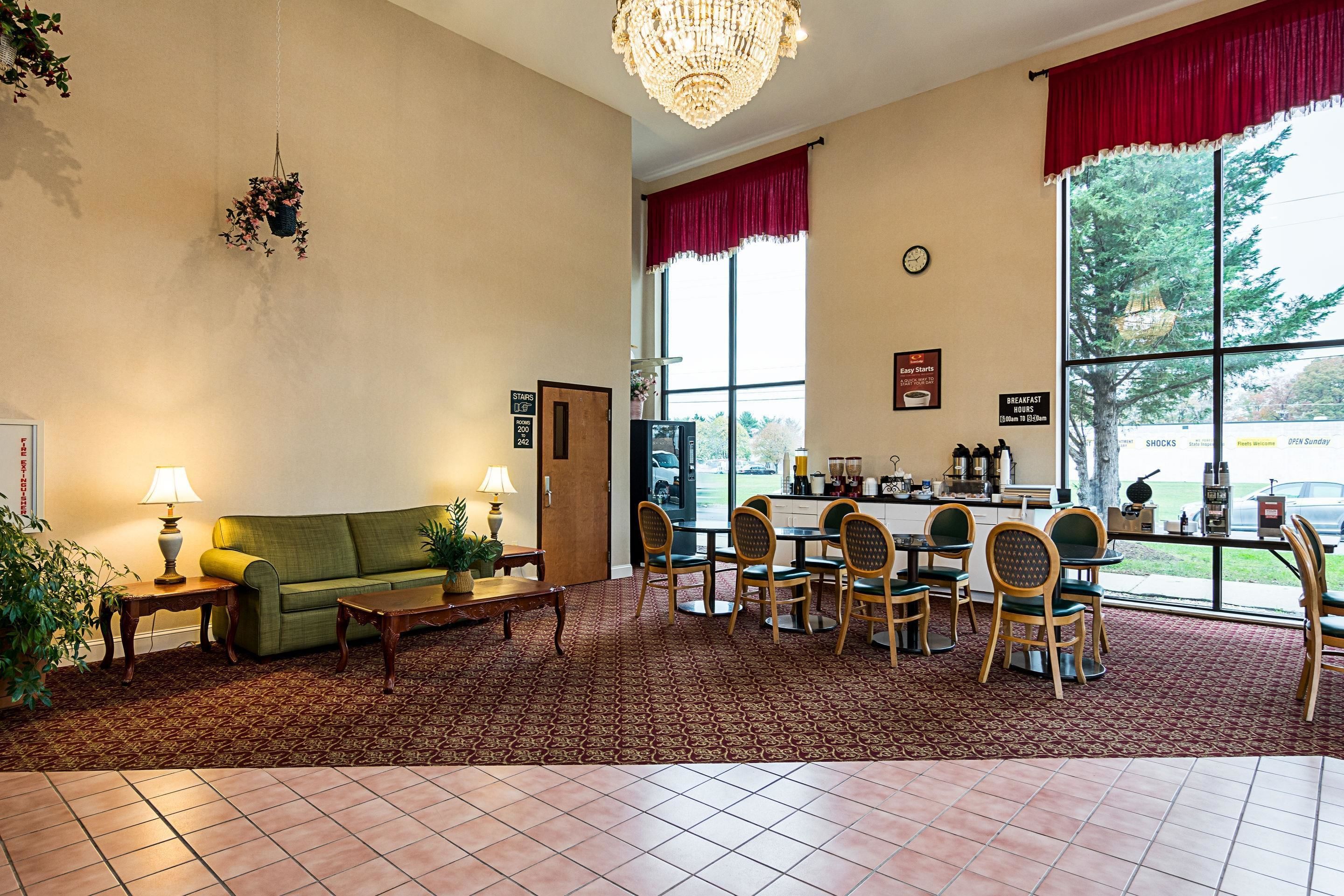A hotel lobby with a green sofa, wooden chairs, a coffee station, and a large window with a view of trees.