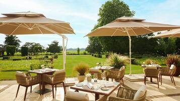 An outdoor dining area with tables and chairs under umbrellas, surrounded by a lush garden and a view of the countryside.