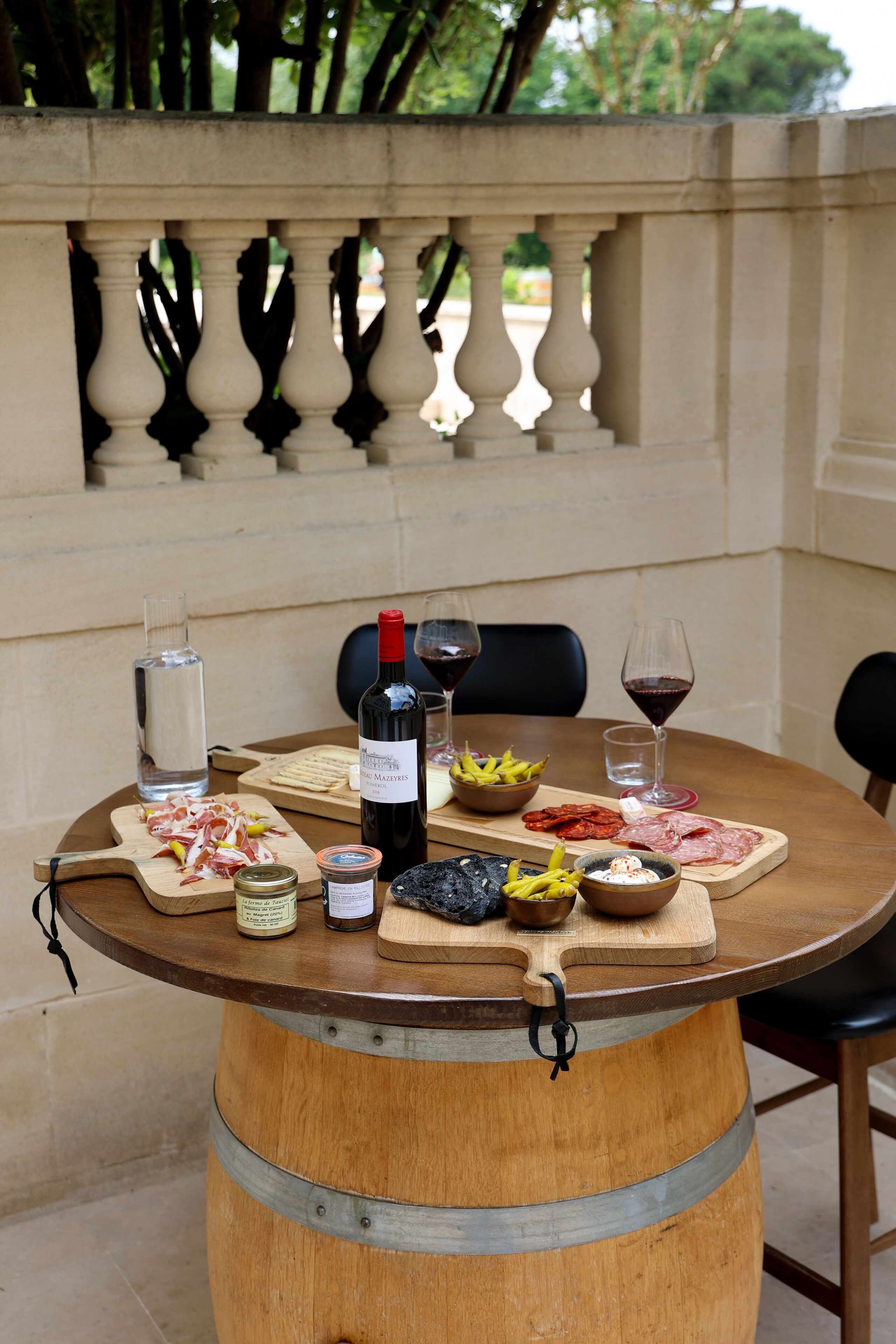 A round wooden table with a barrel base, set outdoors with a view of a stone balustrade and greenery.