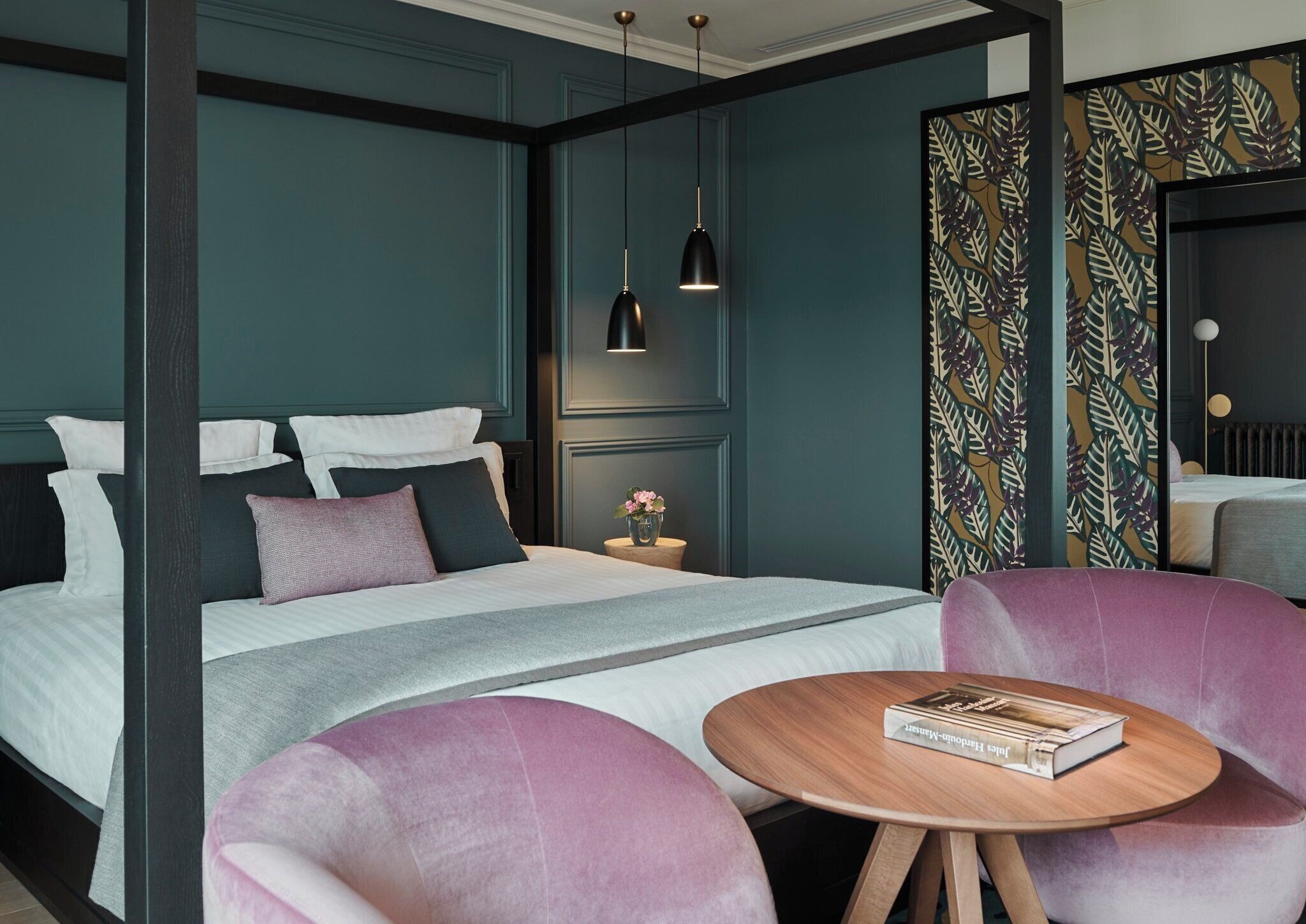 A modern hotel room with a large bed, a bedside table with a vase of flowers, a round wooden table with a book, and a patterned curtain.