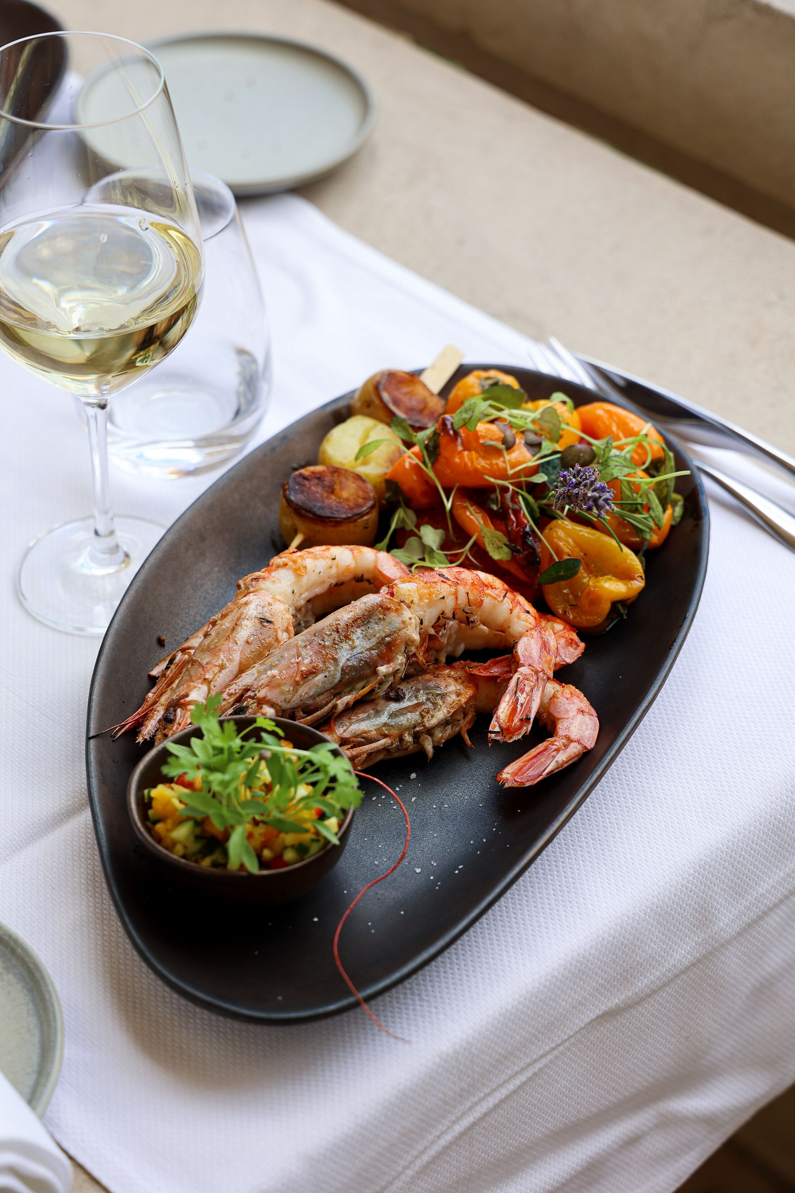 Um prato de comida com camarões e vegetais, um copo de vinho branco e uma pequena tigela de molho.