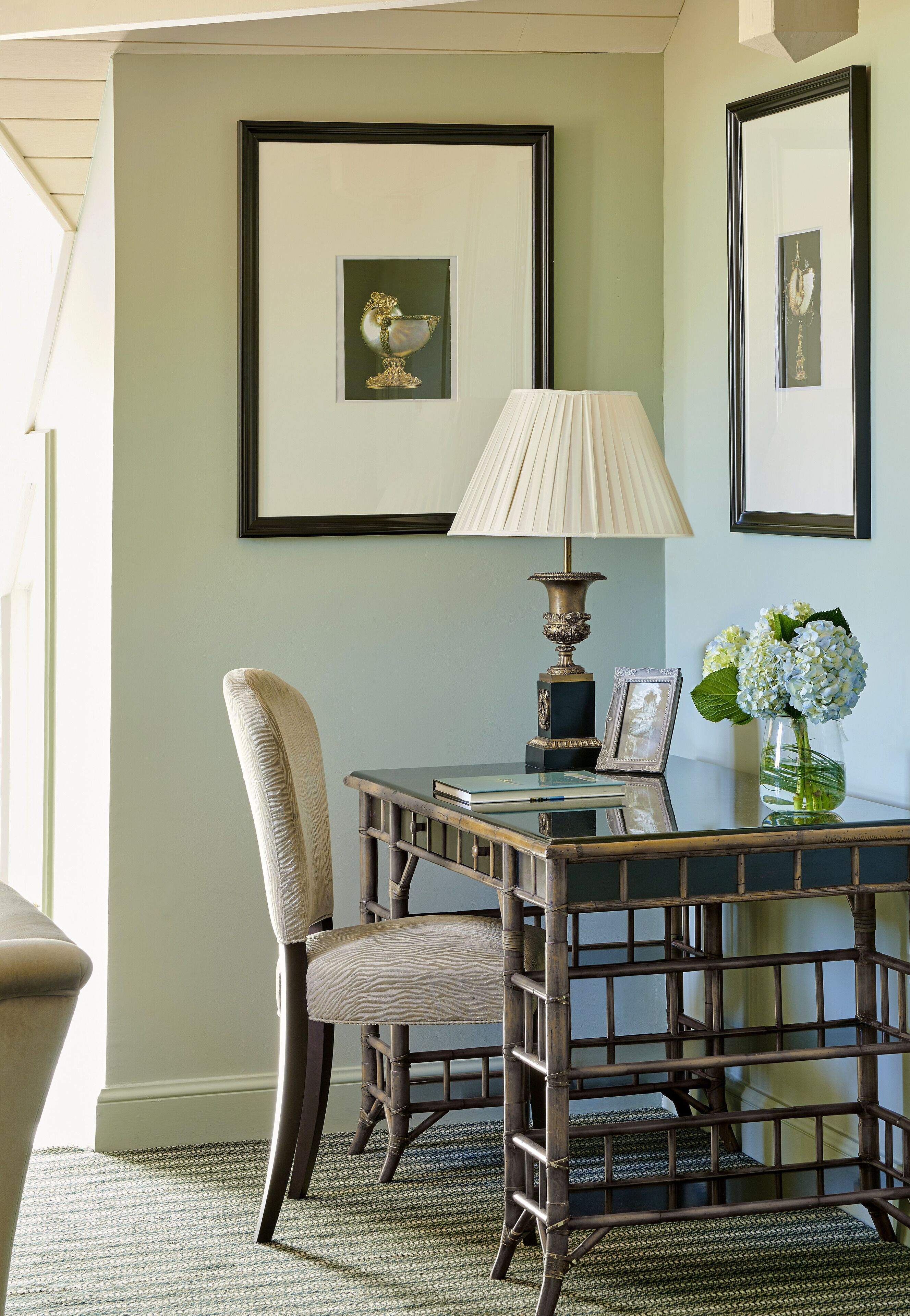 A room with a wooden desk, a chair, a lamp, framed pictures, and a vase with flowers.