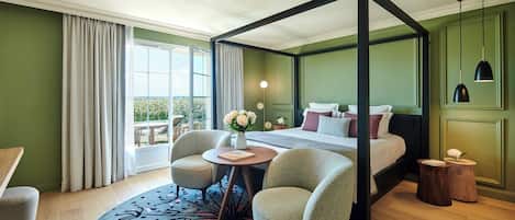 Una cama con dosel con vista a un jardín, una mesa redonda con un jarrón de flores y dos sillones en una habitación con paredes verdes y suelo de madera.