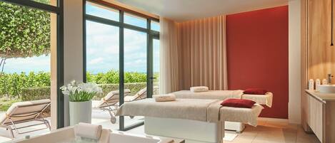 A spa room with a large window, two massage beds, a bathtub, and a sink.