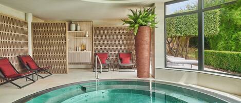 A modern indoor swimming pool with a glass wall, red lounge chairs, and a large window overlooking greenery.