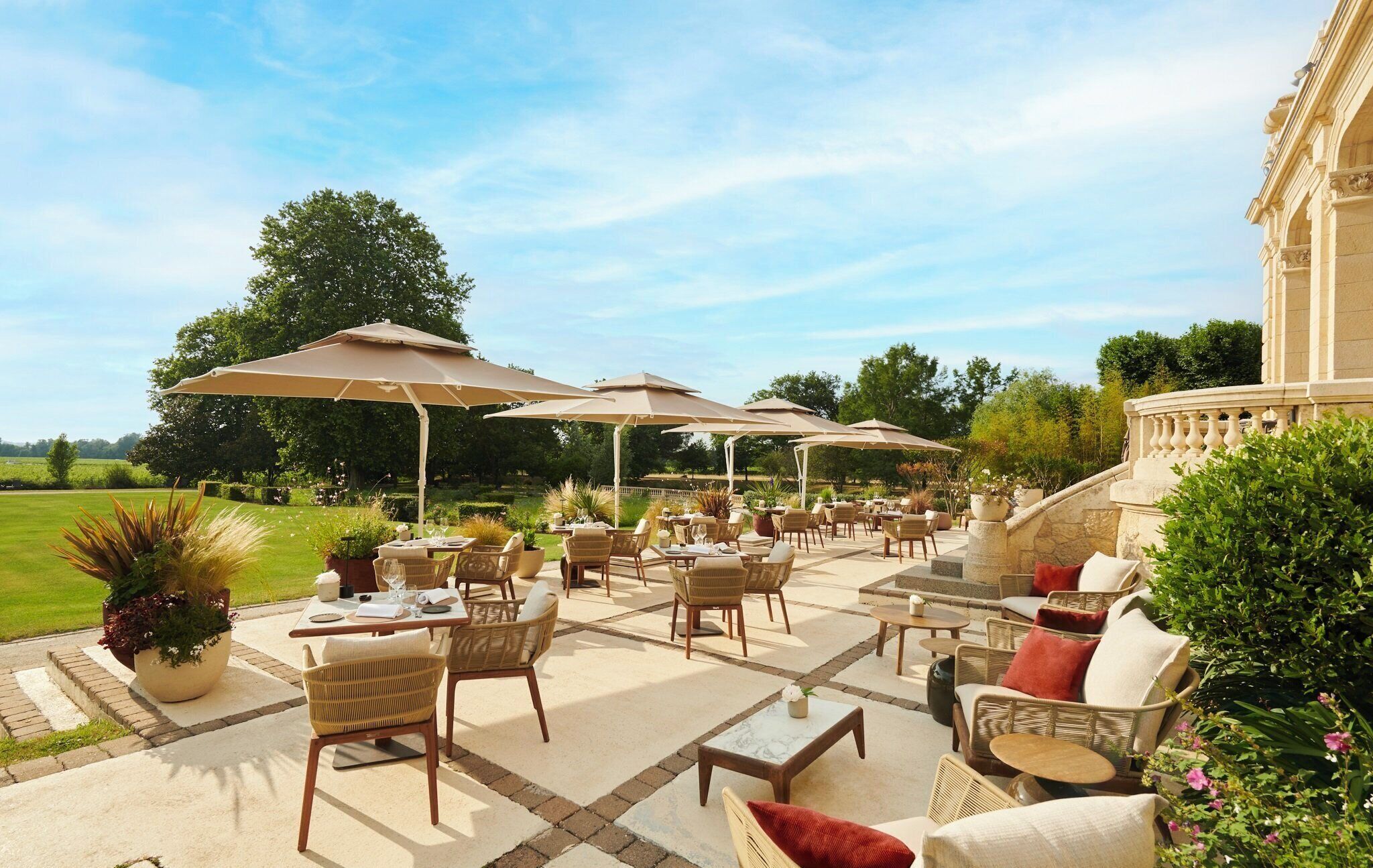 An outdoor patio with wicker furniture, beige umbrellas, and a stone building in the background.