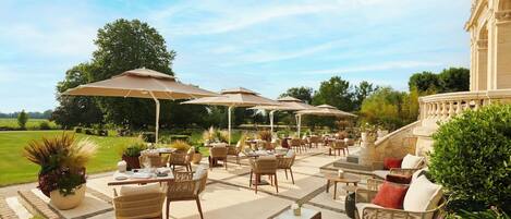 An outdoor patio with wicker furniture, beige umbrellas, and a stone building in the background.
