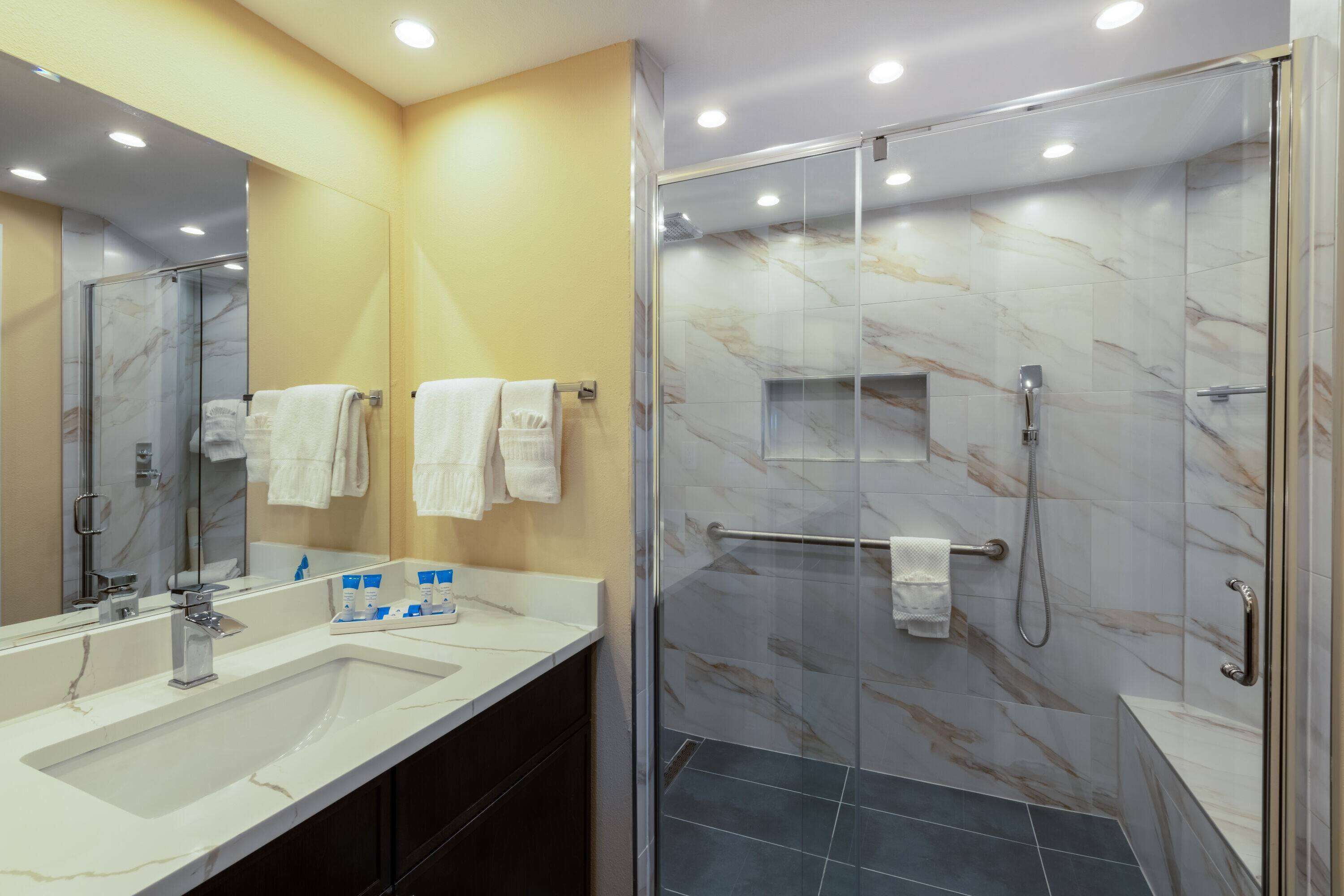 Een moderne badkamer met een grote spiegel, twee wastafels, een douche met glazen deuren en marmeren wanden.