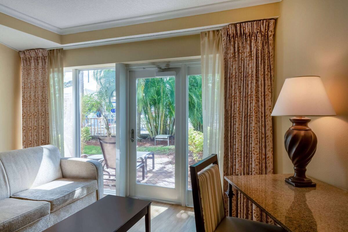 A hotel room with a sofa, a table, a lamp, and a sliding glass door leading to an outdoor area with a patio and greenery.