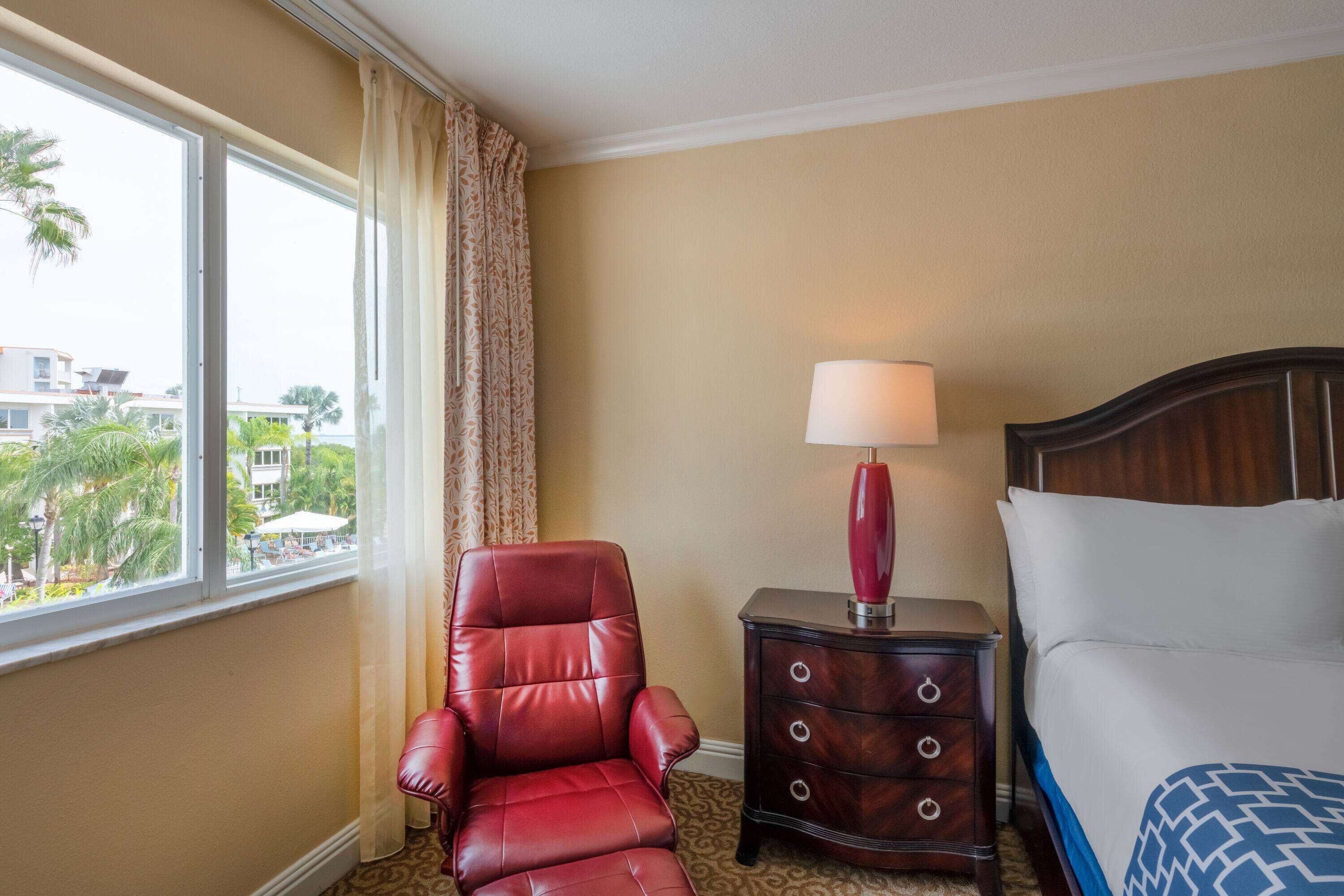 A hotel room with a bed, a red leather chair, a nightstand with a lamp, and a view of palm trees and buildings outside.