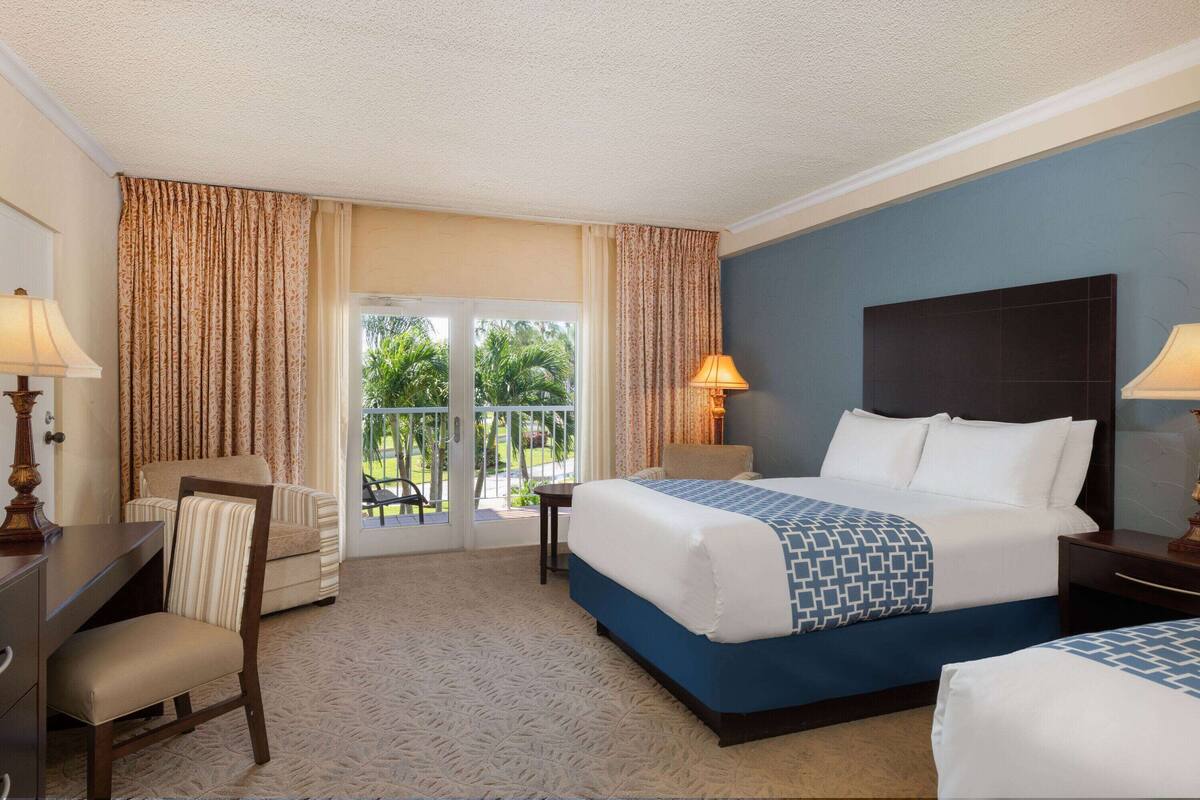A hotel room with two beds, a desk, a chair, a lamp, and a view of greenery through the door.