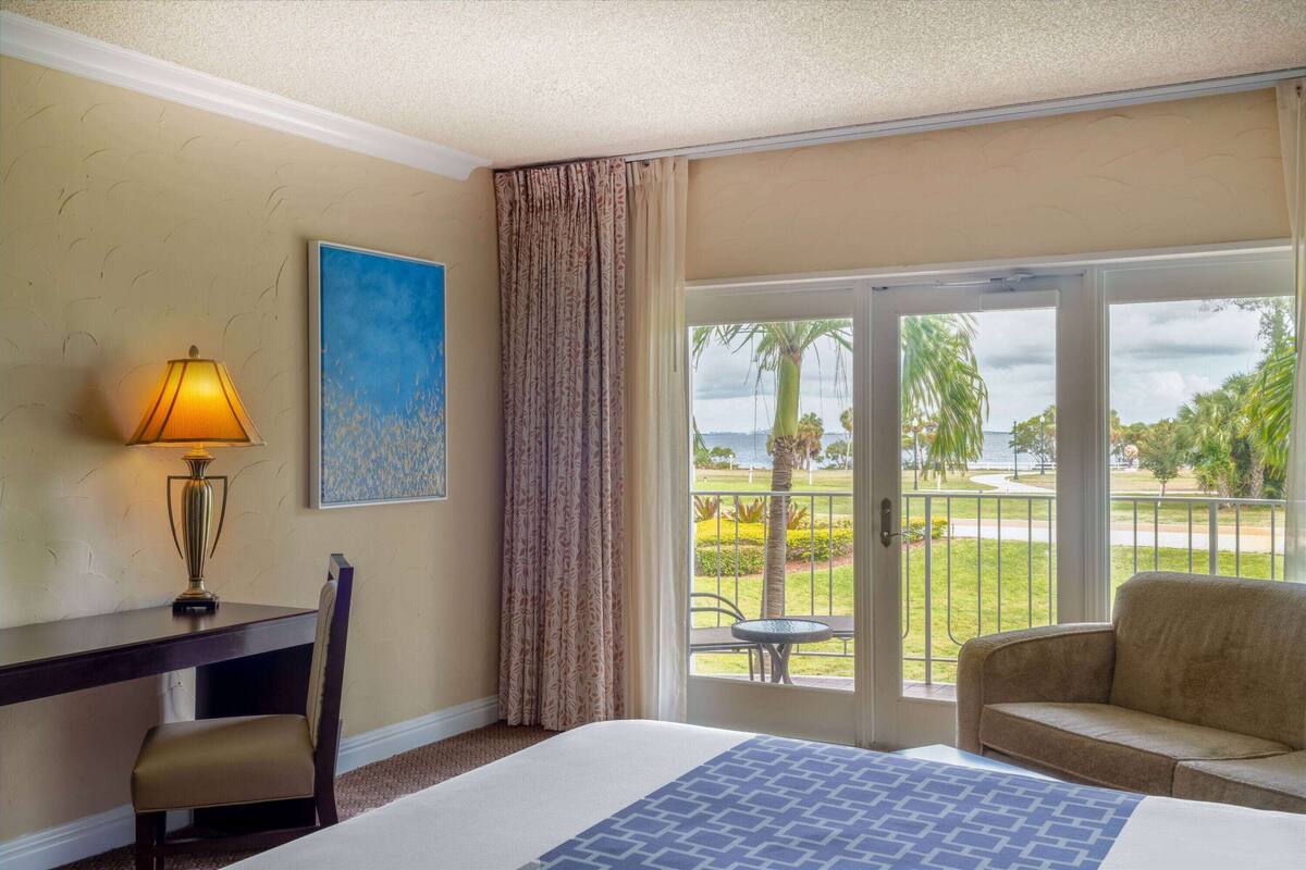 A hotel room with a bed, a desk with a lamp, a chair, a couch, and a large sliding glass door leading to a balcony with a view of palm trees and a body of water.