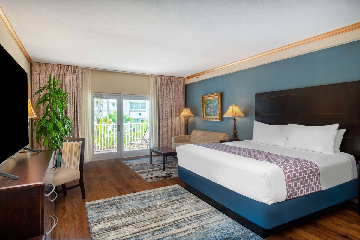 A hotel room with a large bed, a desk, a chair, a plant, and a balcony with a view.