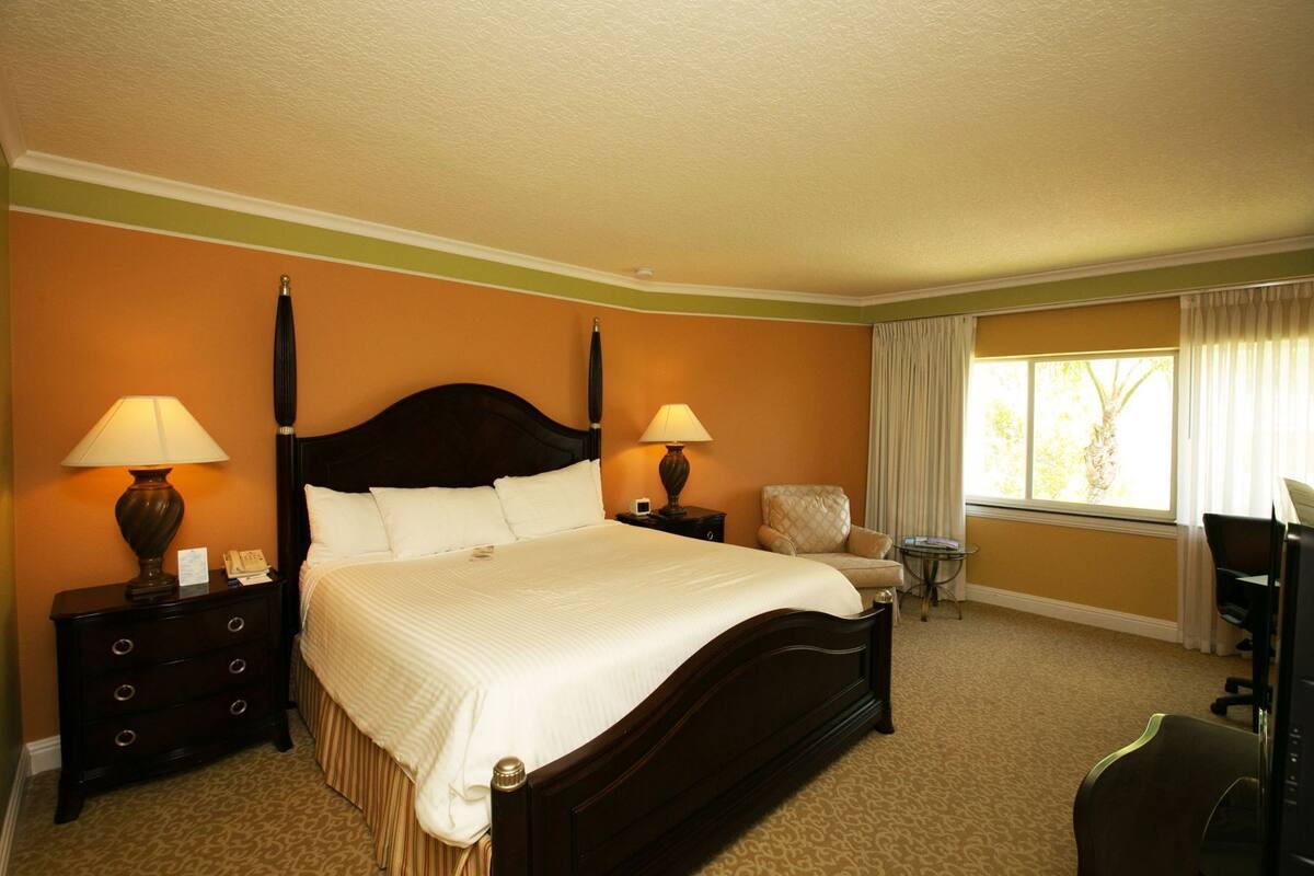A hotel room with a large bed, two bedside tables with lamps, a chair, a small table, and a desk with a chair.