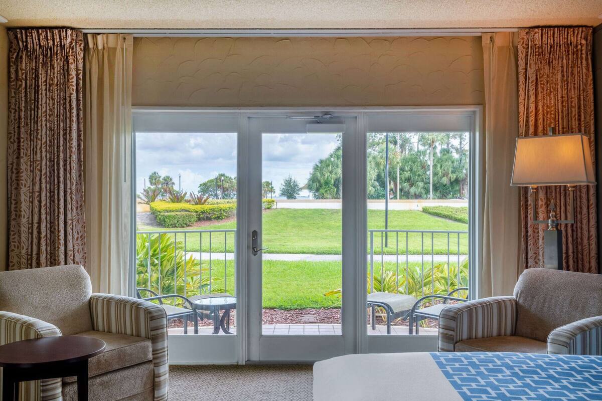 A hotel room with a large sliding glass door leading to a balcony with a view of a landscaped garden and palm trees. There are two armchairs and a small table on the balcony.
