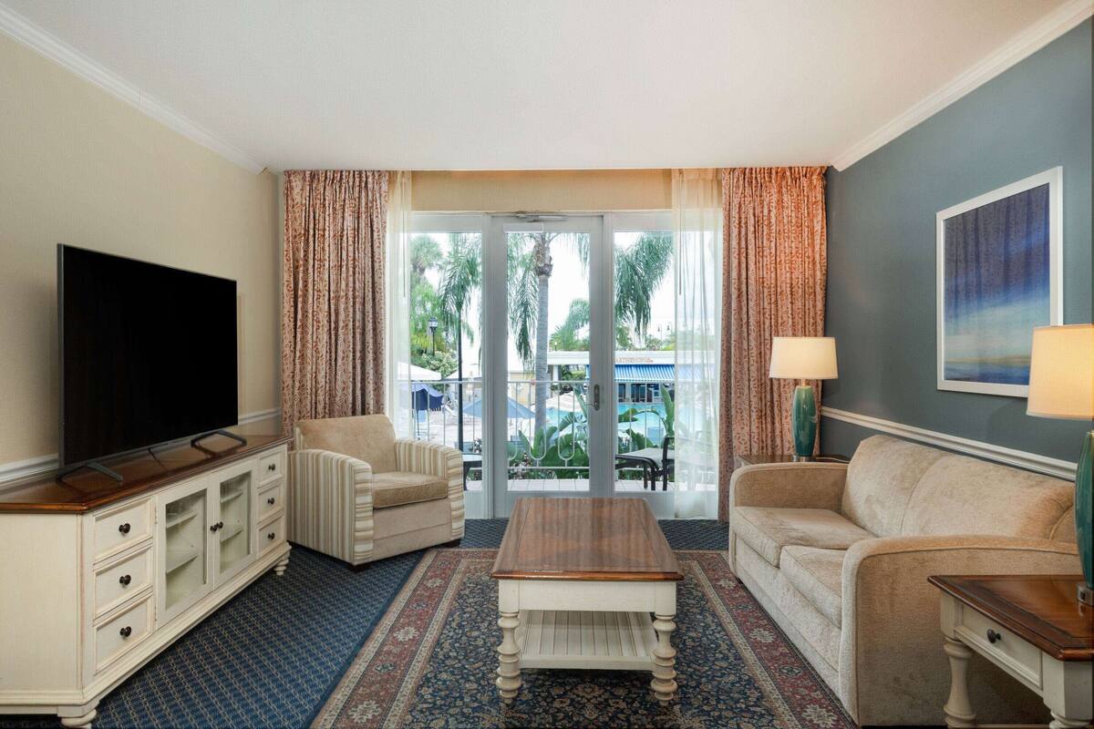 A hotel room with a television, two sofas, a coffee table, and a view of palm trees through the glass door.