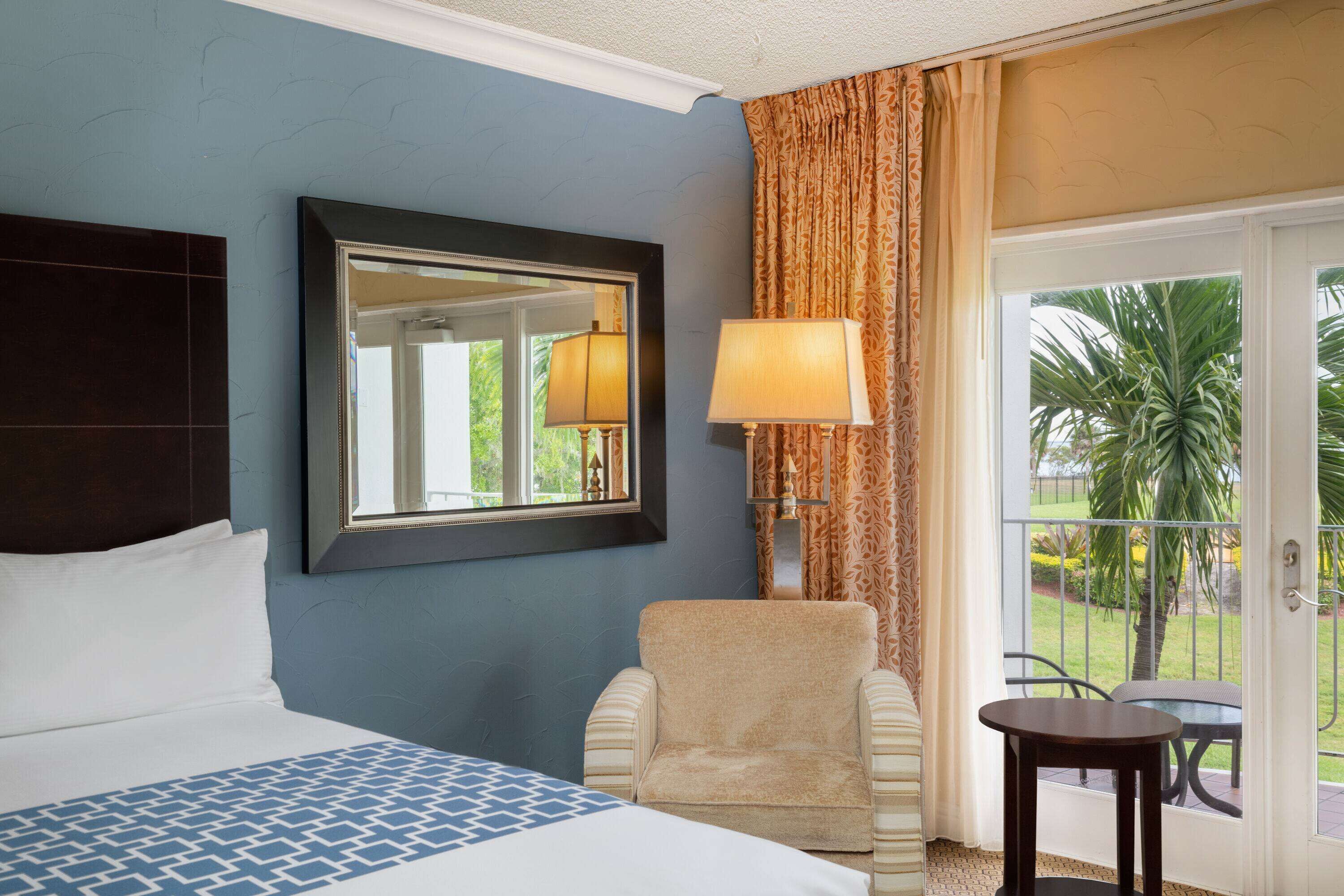 A hotel room with a bed, a chair, a table, a lamp, a mirror, and a view of the outdoors.