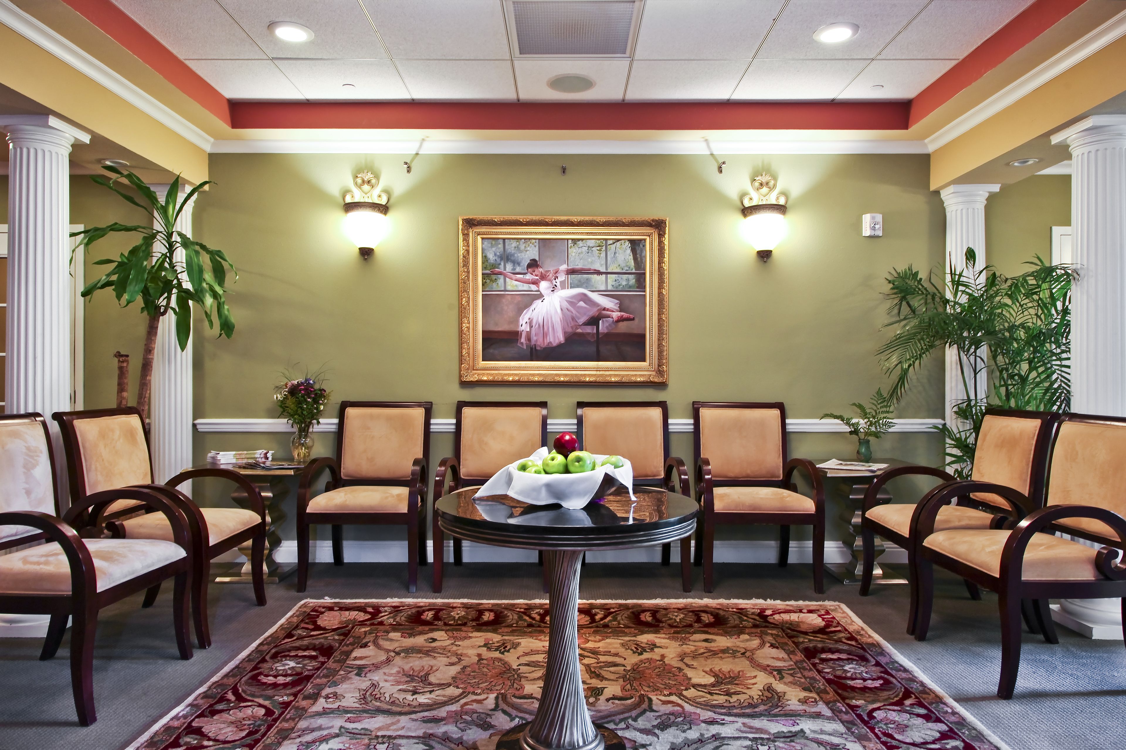 A lobby with a round table, chairs, a painting, and a vase with apples.