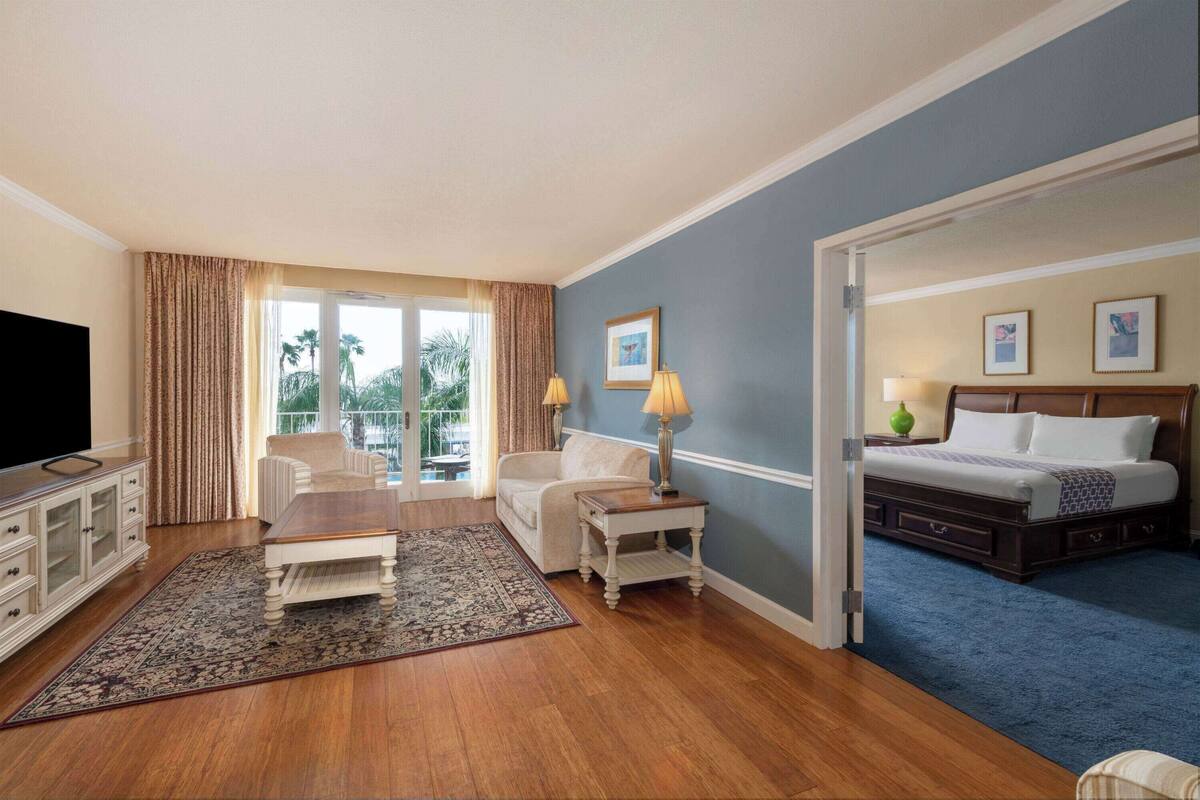 A hotel room with a wooden floor, a sofa, a coffee table, a TV, a bed with a blue carpet, and a balcony with curtains.