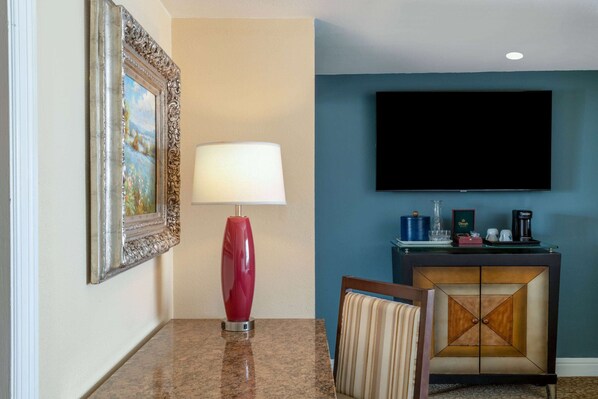 Una habitación de hotel con una mesa de comedor, una lámpara roja, un cuadro enmarcado, un televisor y un minibar.