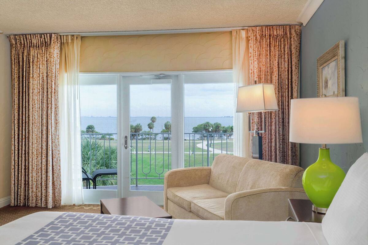 A hotel room with a bed, a beige sofa, a green lamp, and a view of the sea through a large window.
