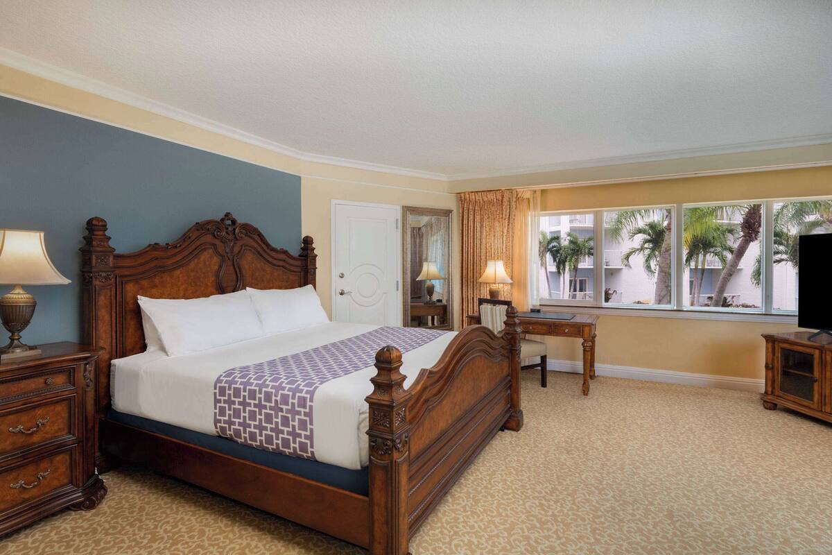 A hotel room with a large bed, a desk, a chair, a television, and a window with a view of palm trees.