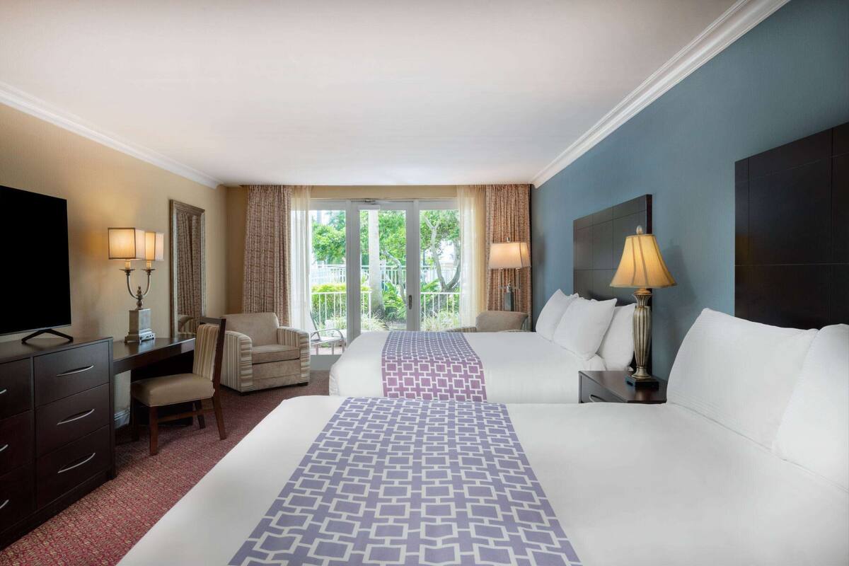 A hotel room with two beds, a desk, a television, and a balcony with curtains.