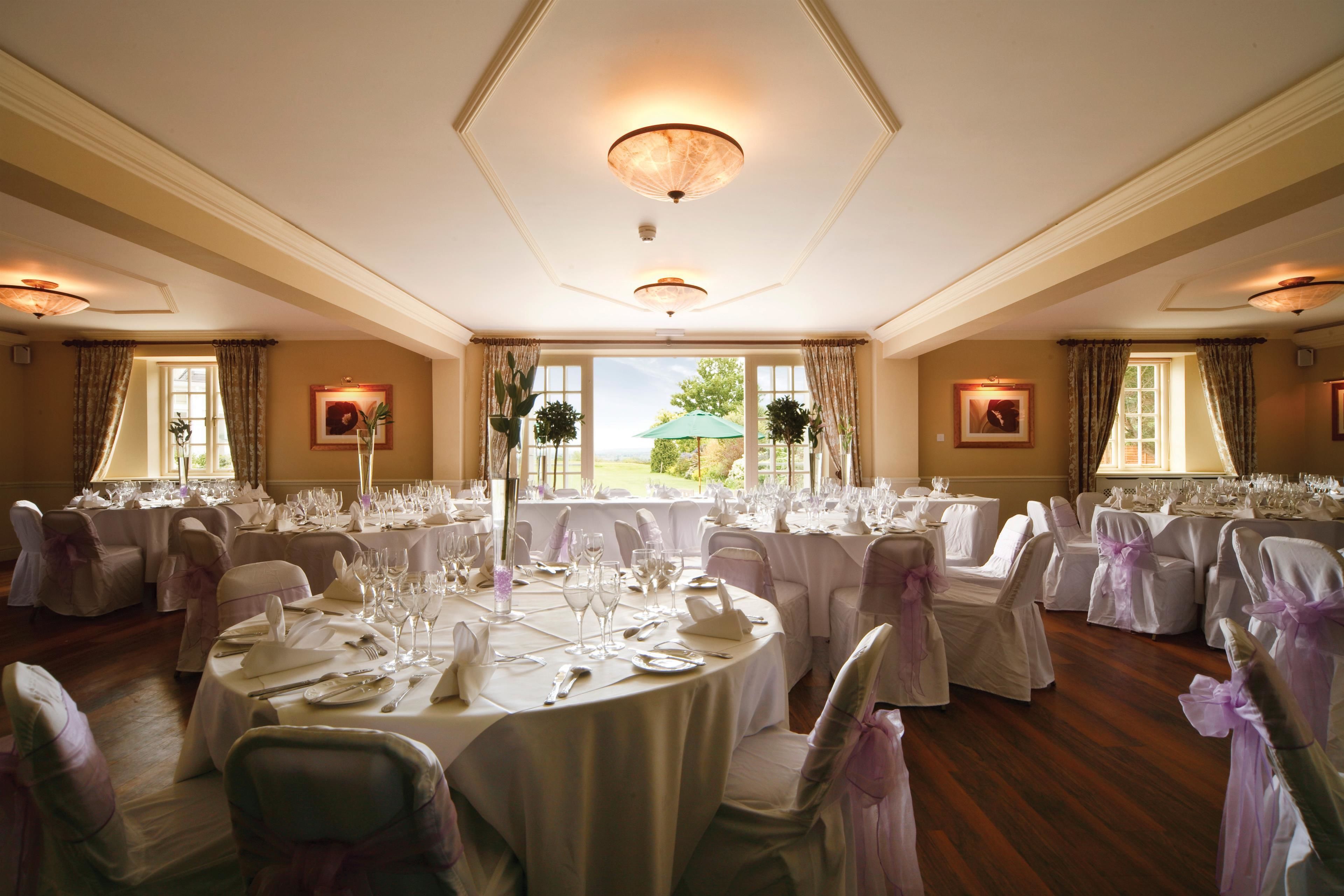 Un salón de banquetes con mesas redondas dispuestas para un evento formal, ventanas amplias con cortinas y vistas al exterior.