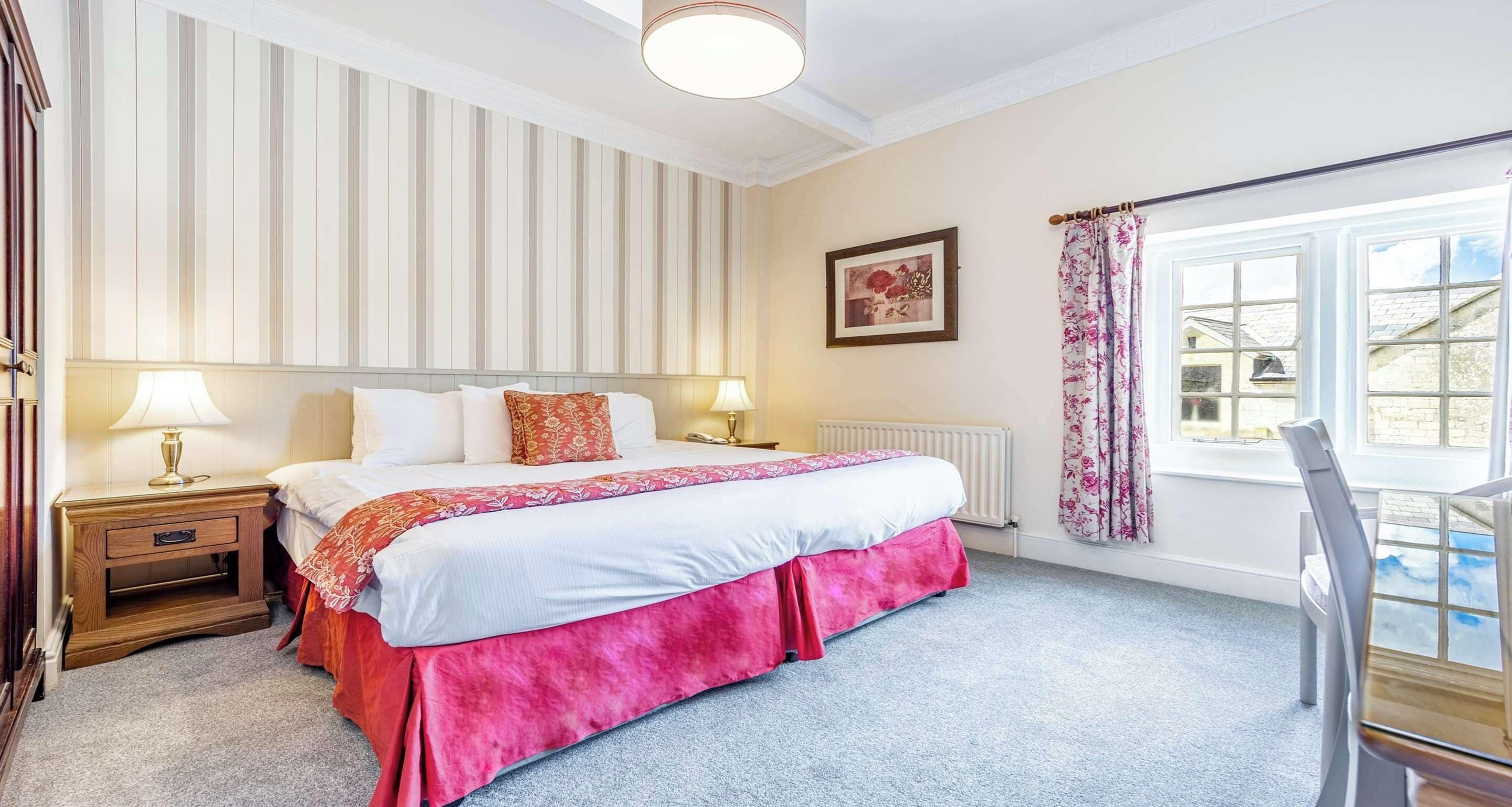 A bedroom with a large bed, two bedside tables with lamps, a radiator, a window with curtains, and a framed picture on the wall.